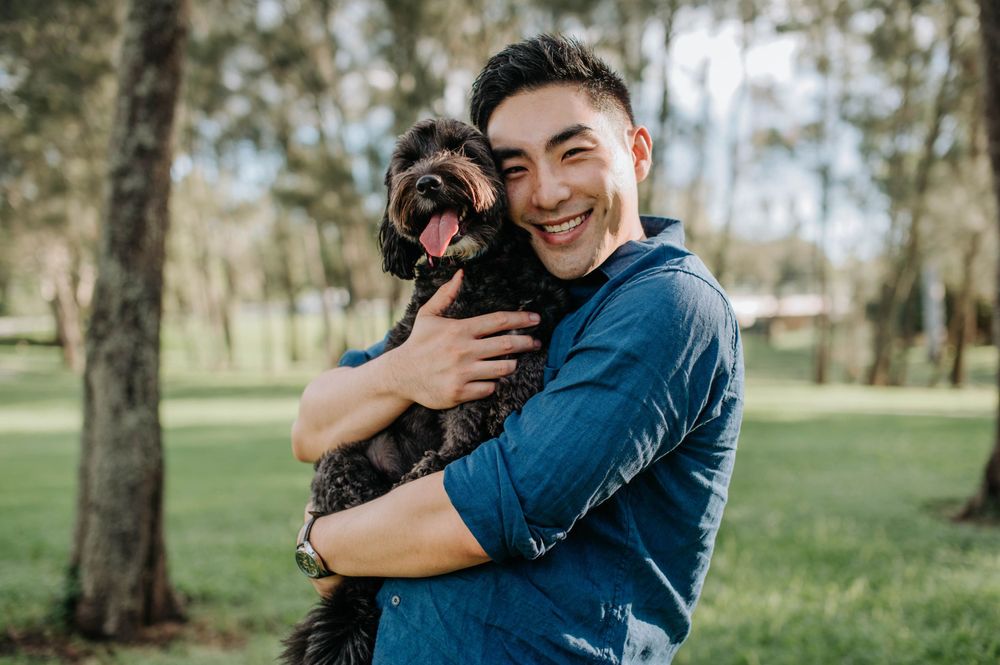 Person in blue shirt joyfully hugs black dog in sunny park setting.
