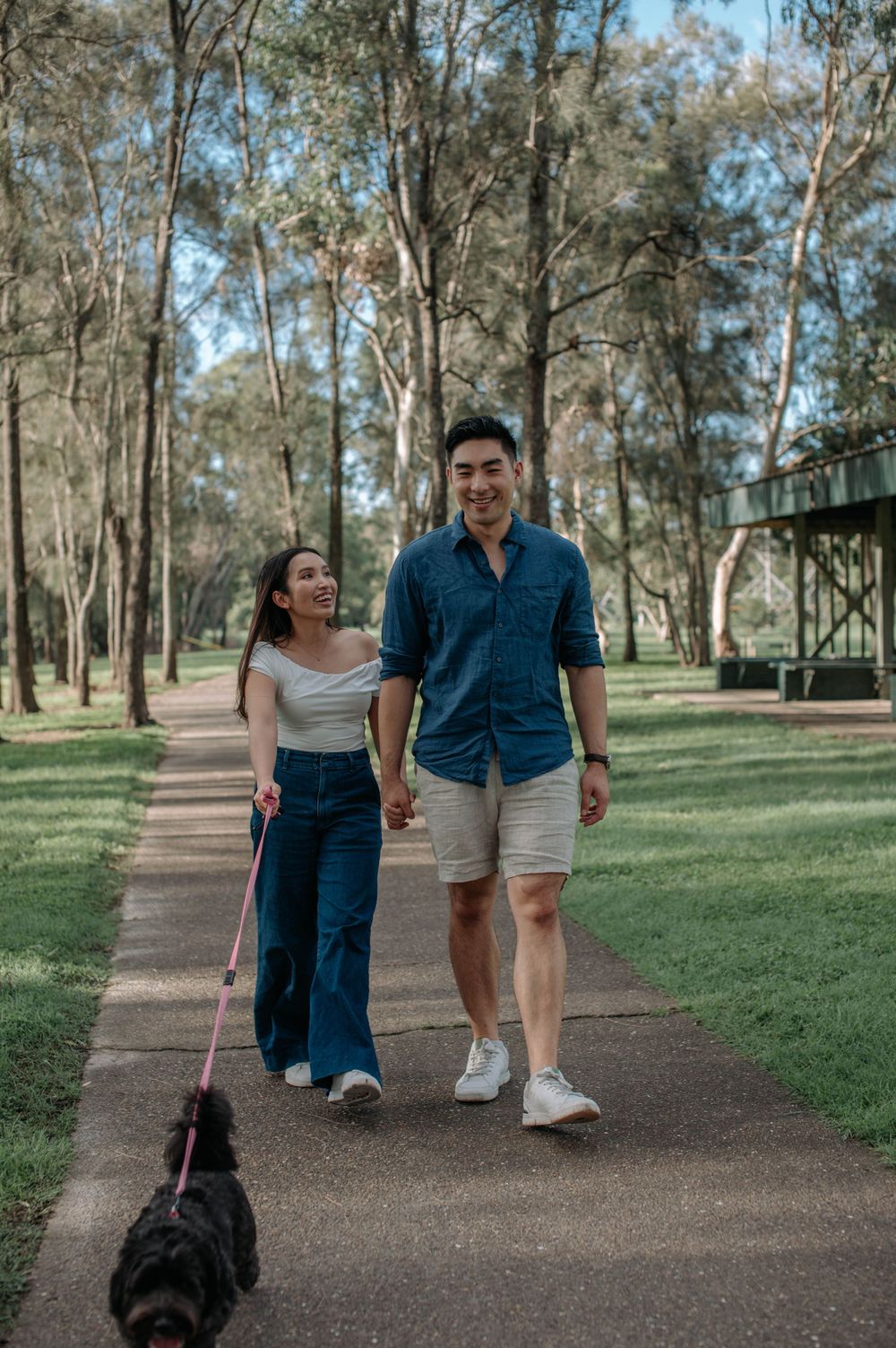 Couple walks their black dog along a paved path through a peaceful wooded park.