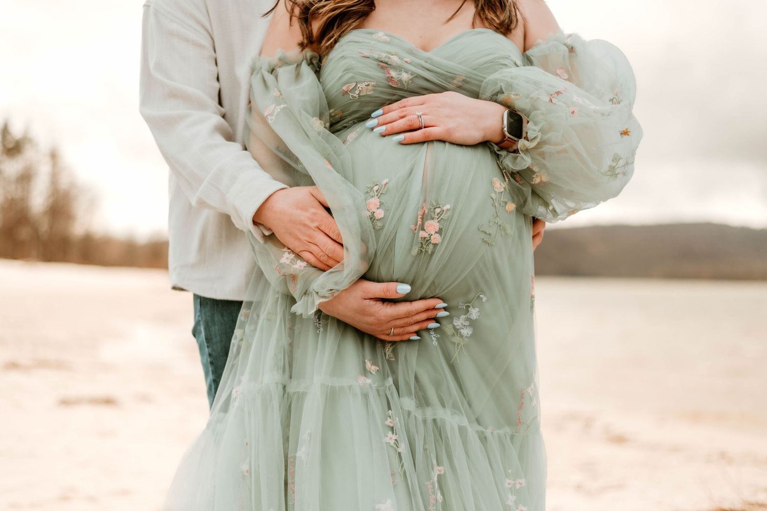 Maternity photo showing couple embracing in flowing mint green dress by waterside.