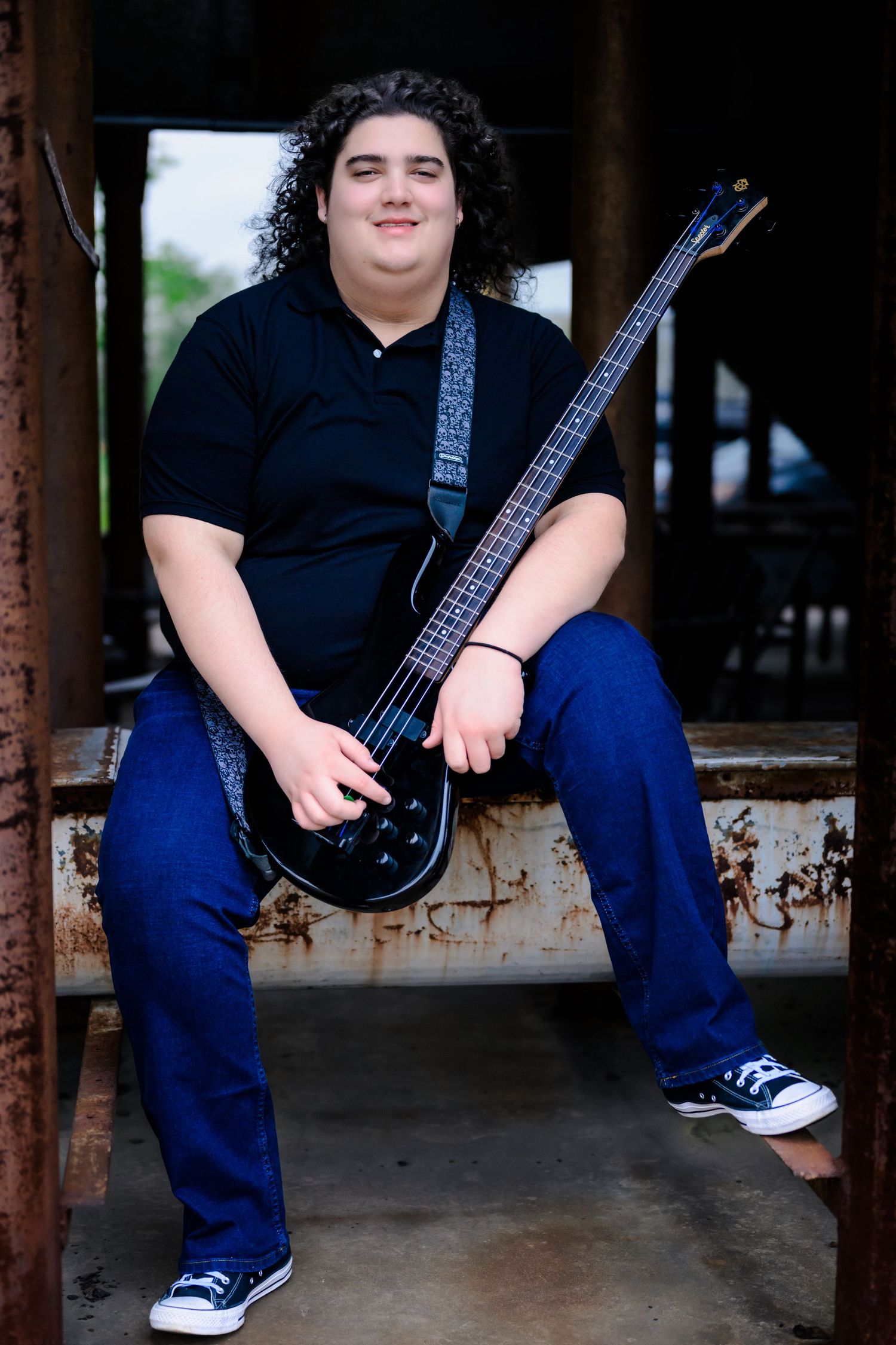 A musician sits with a black bass guitar wearing a navy shirt, blue jeans and sneakers in a rustic setting.