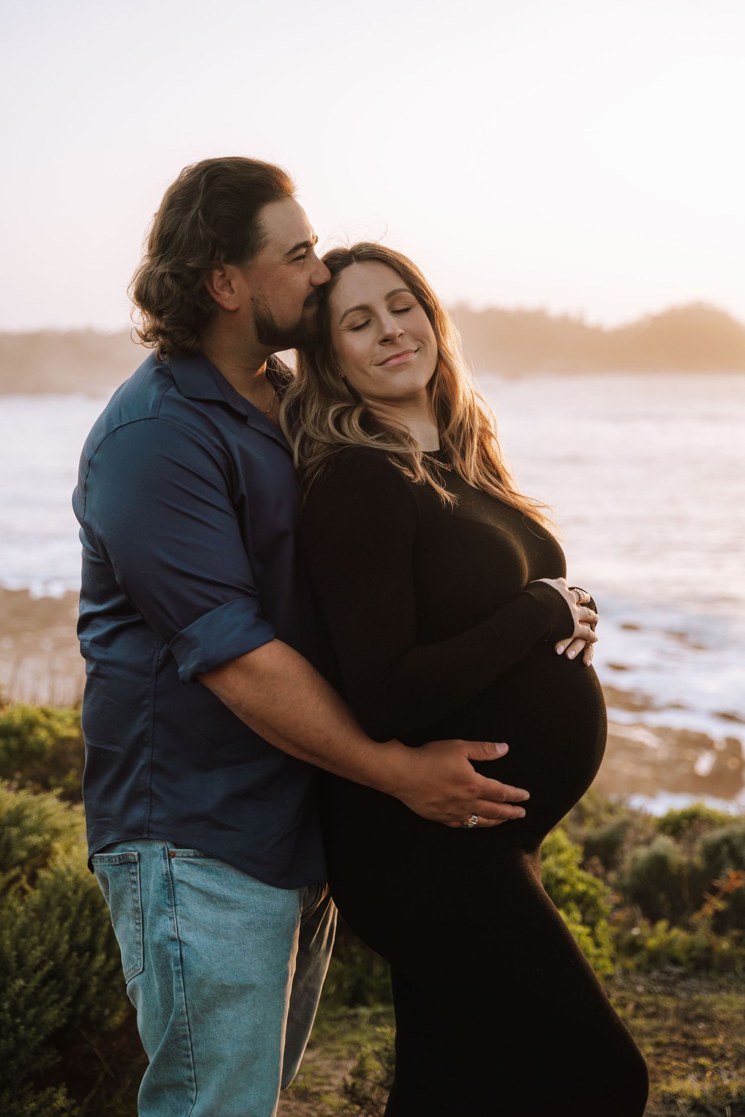 A couple embraces at sunset by the ocean as they take maternity photos on a coastal cliff.