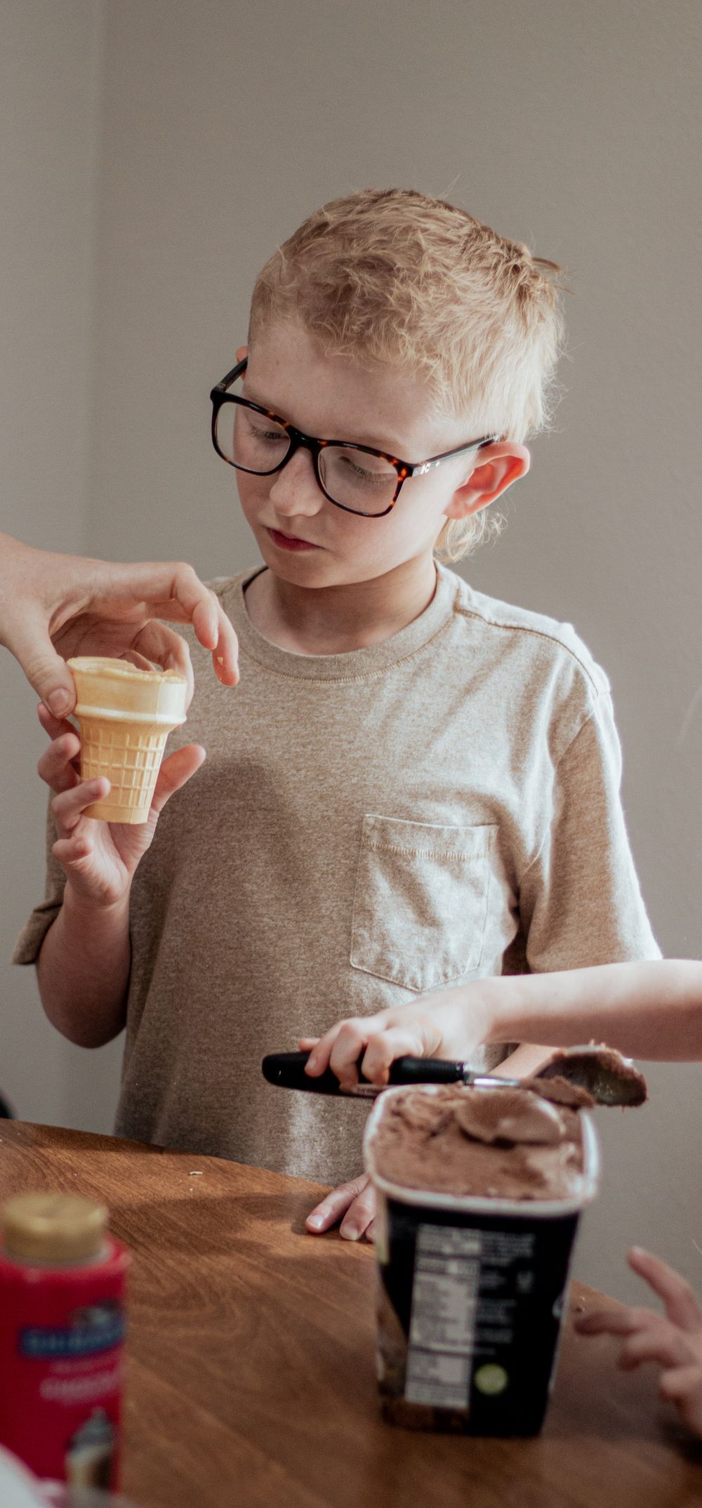 Child in glasses examining an ice cream cone and container of ice cream on wooden table.