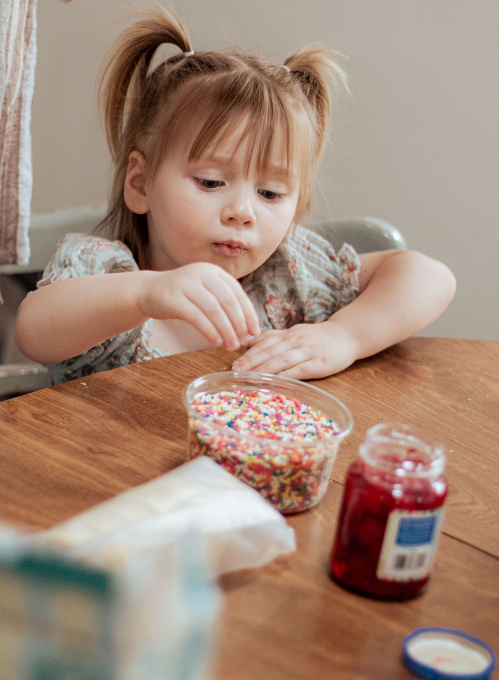Young child in pigtails decorating treats with colorful sprinkles at a wooden table.