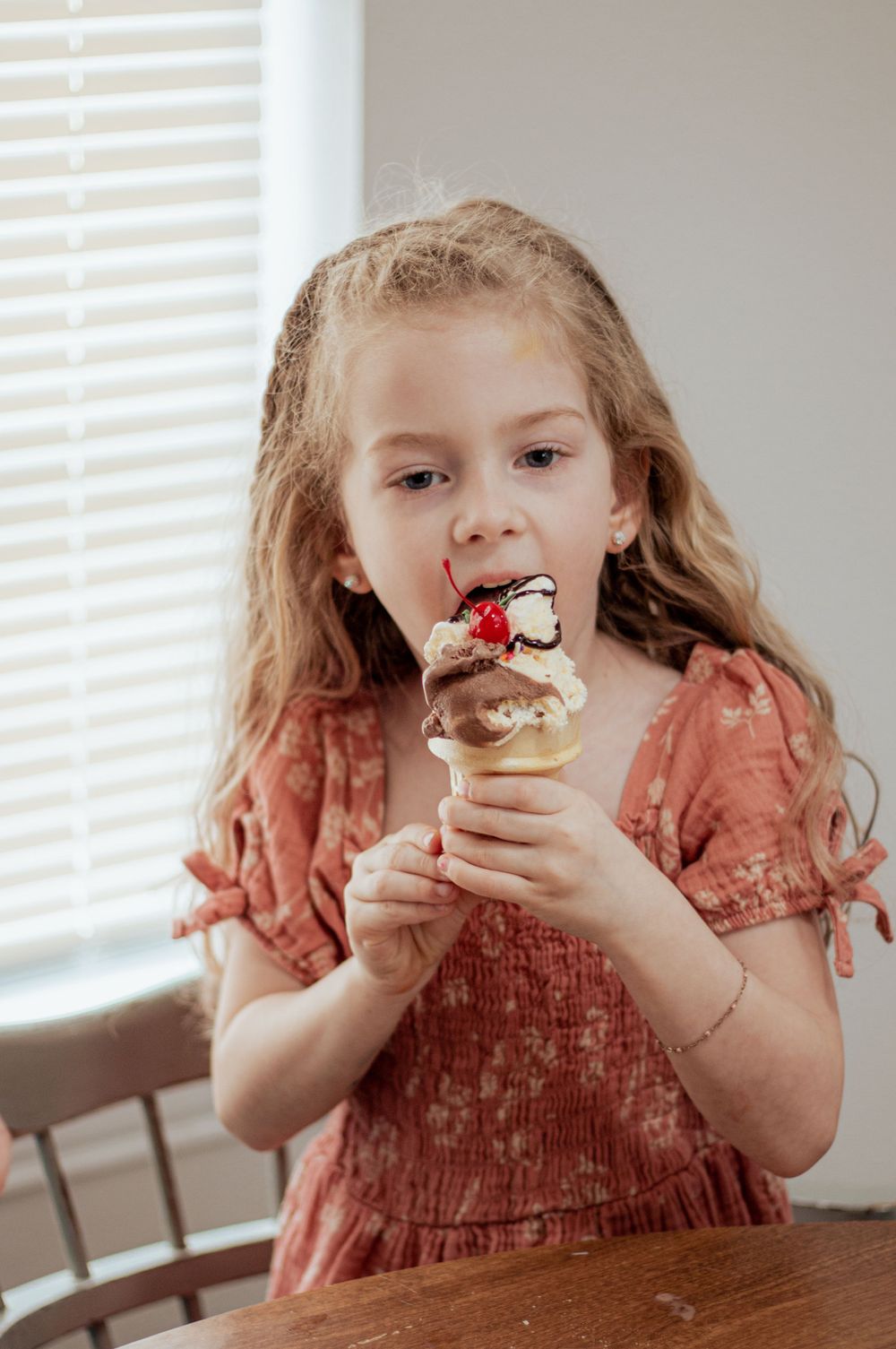 A young child in a pink dress enjoying an ice cream cone with whipped cream and chocolate sauce by a window.