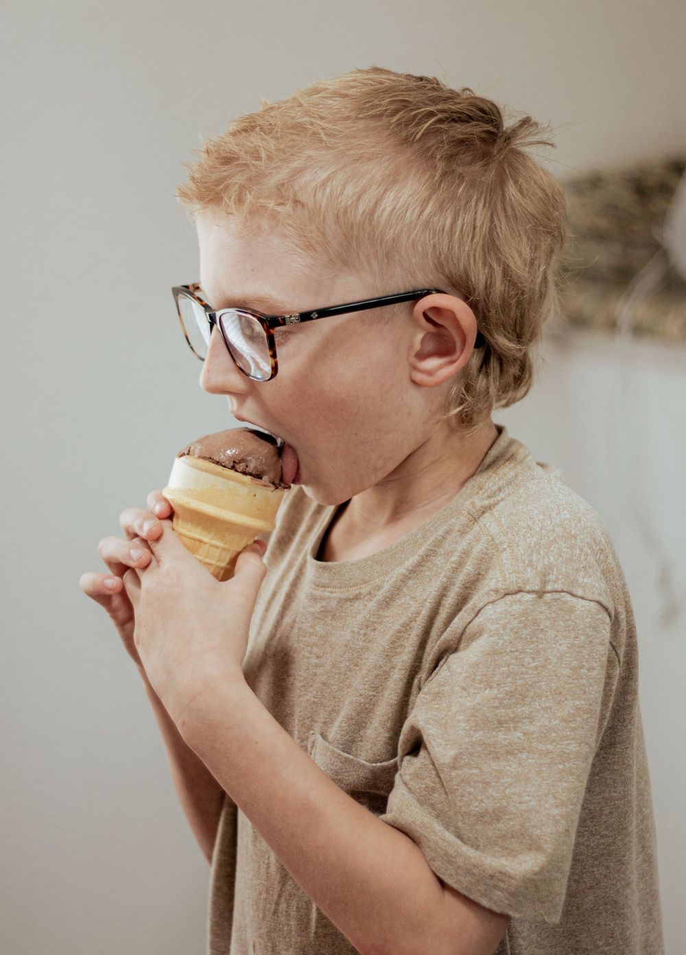 Child wearing glasses takes a big bite of an ice cream treat while wearing a beige shirt.