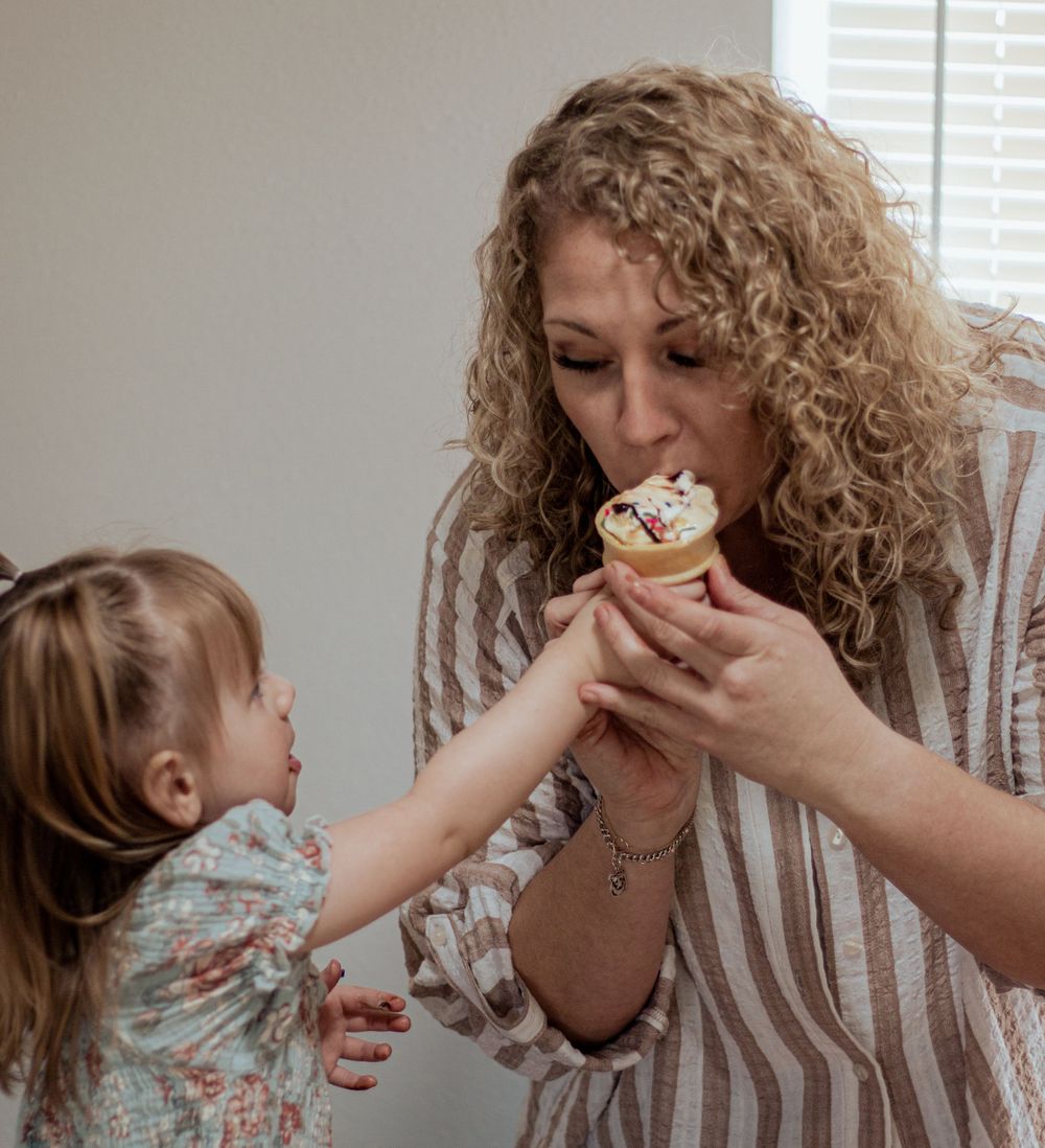 A person with curly blonde hair eating a cupcake while a small child reaches up to share the treat.