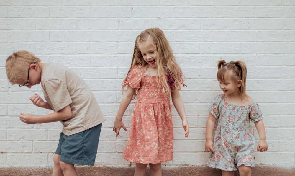Three children in casual sundresses play and dance against a white brick wall background.