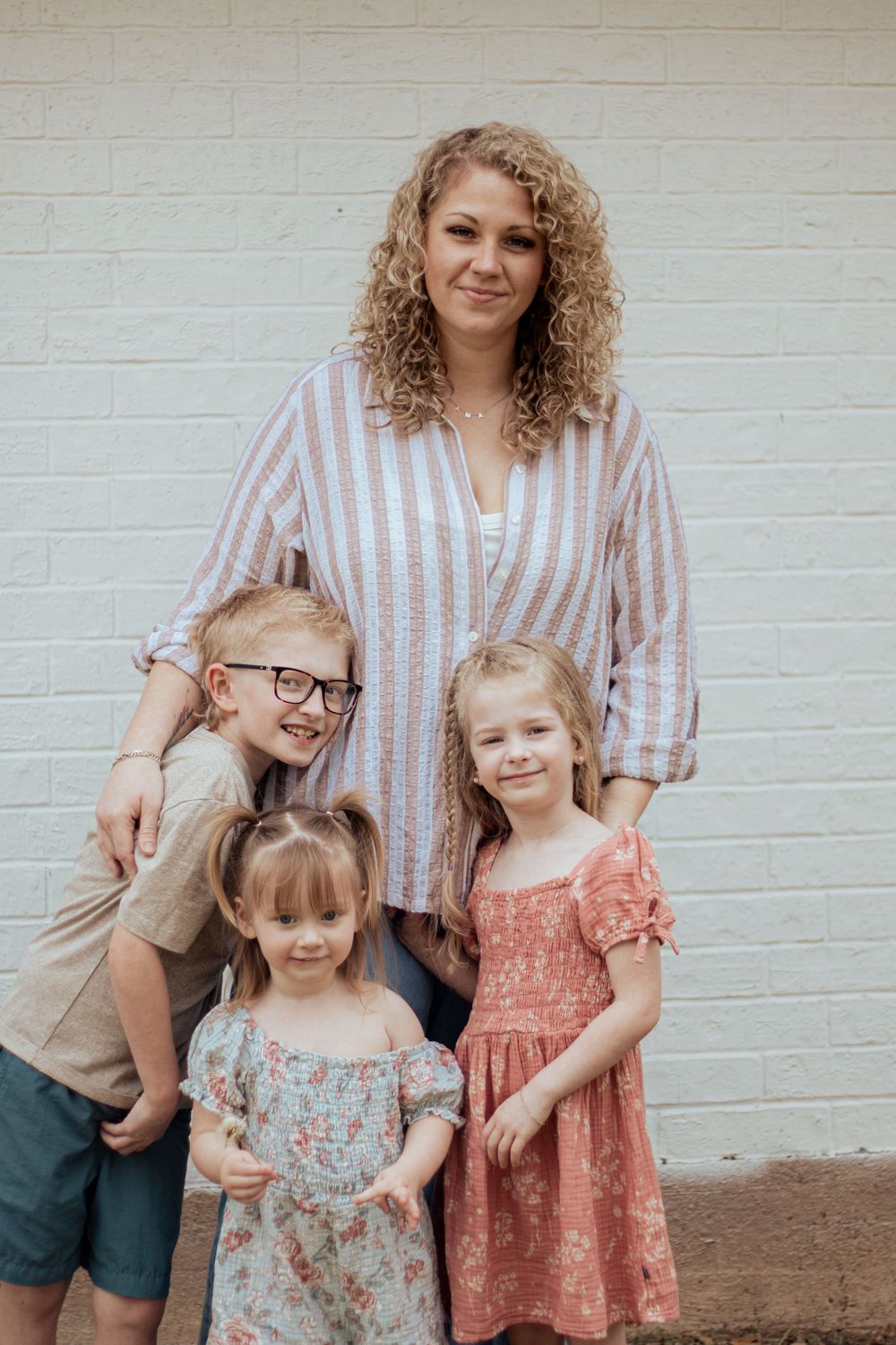 A candid family portrait against a white brick wall featuring a parent with curly hair and three children in summer outfits.