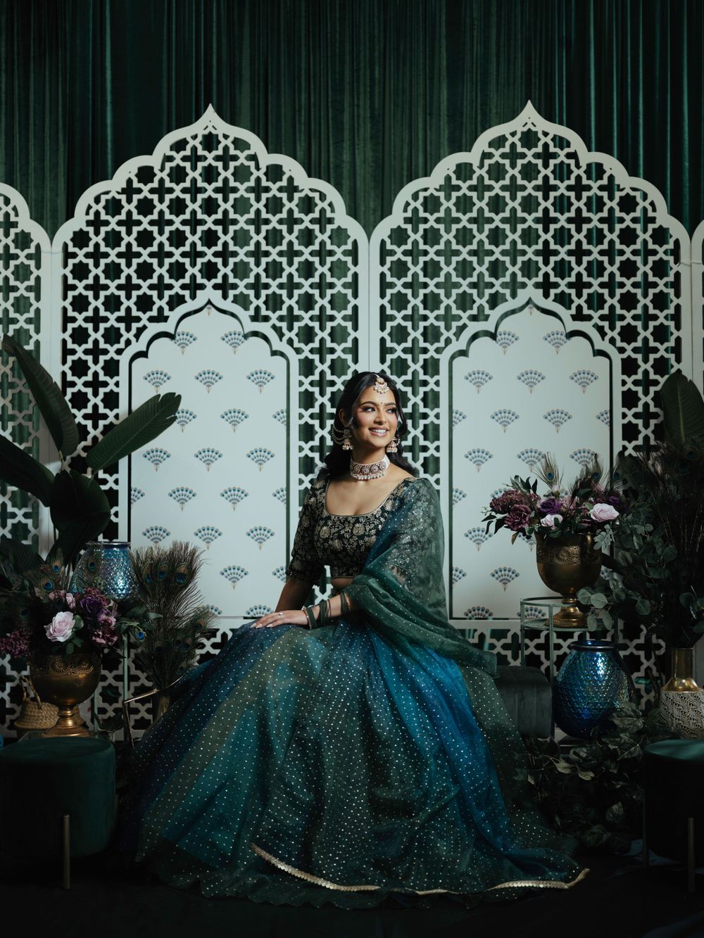 A regal emerald gown adorns a figure posed against ornate Moroccan-style white lattice panels with floral arrangements.
