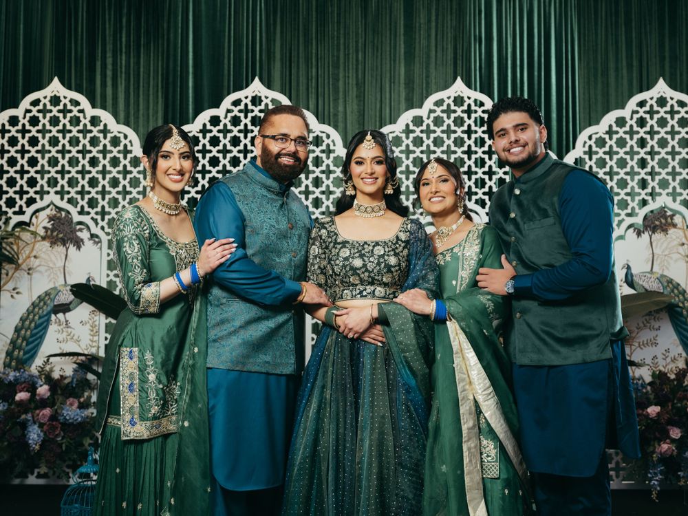 Group photo at an Indian wedding celebration with guests wearing traditional green and blue formal attire against ornate backdrop.