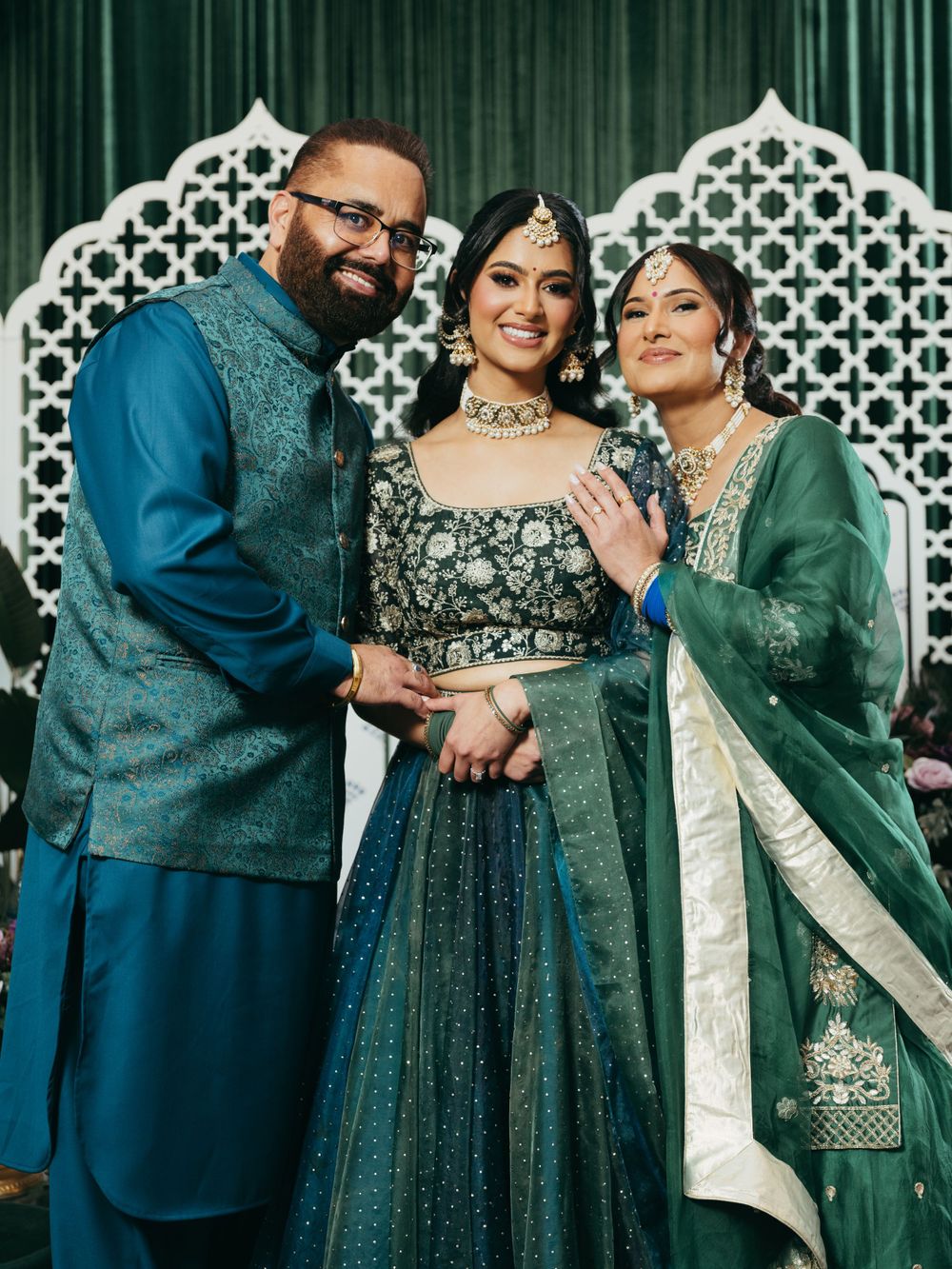 Three people in traditional Indian formal wear pose together against a decorative white lattice backdrop at a celebration.