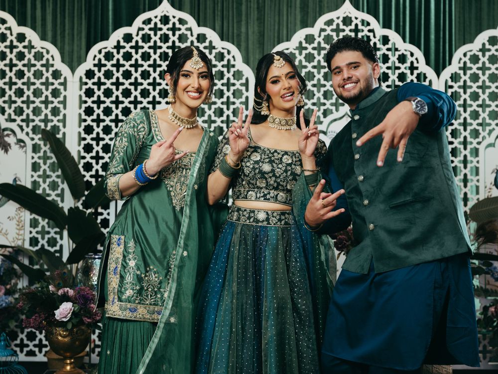Three people in traditional Indian wedding attire posing and laughing together in front of a decorative latticed backdrop.