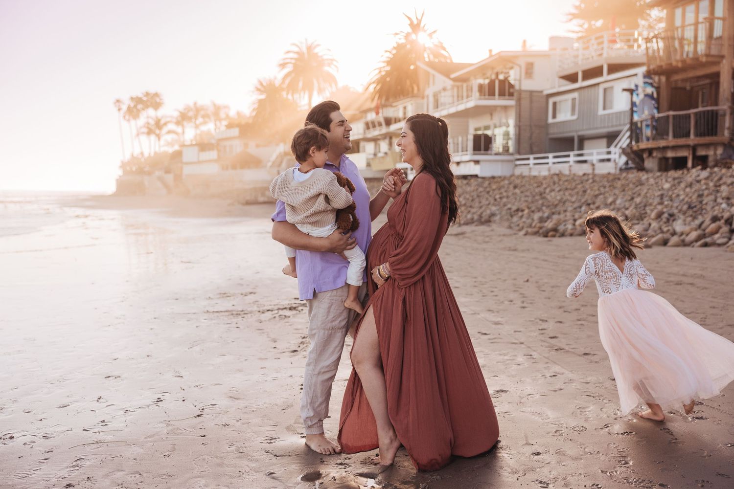 A family enjoys a sunset moment on a sandy beach with houses visible along the coastline in the background.