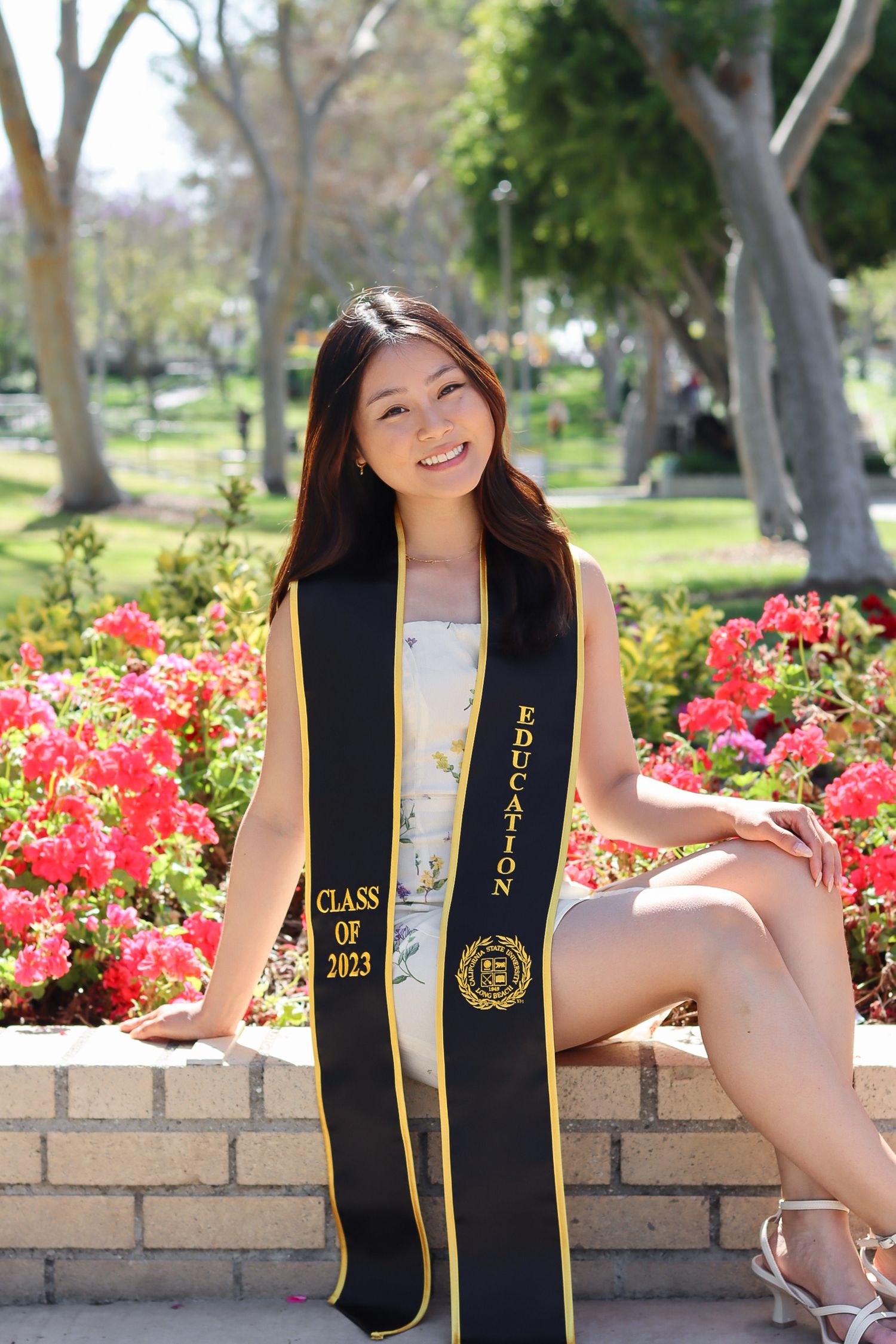 Graduate in white dress with black and gold sash poses by flowering garden.