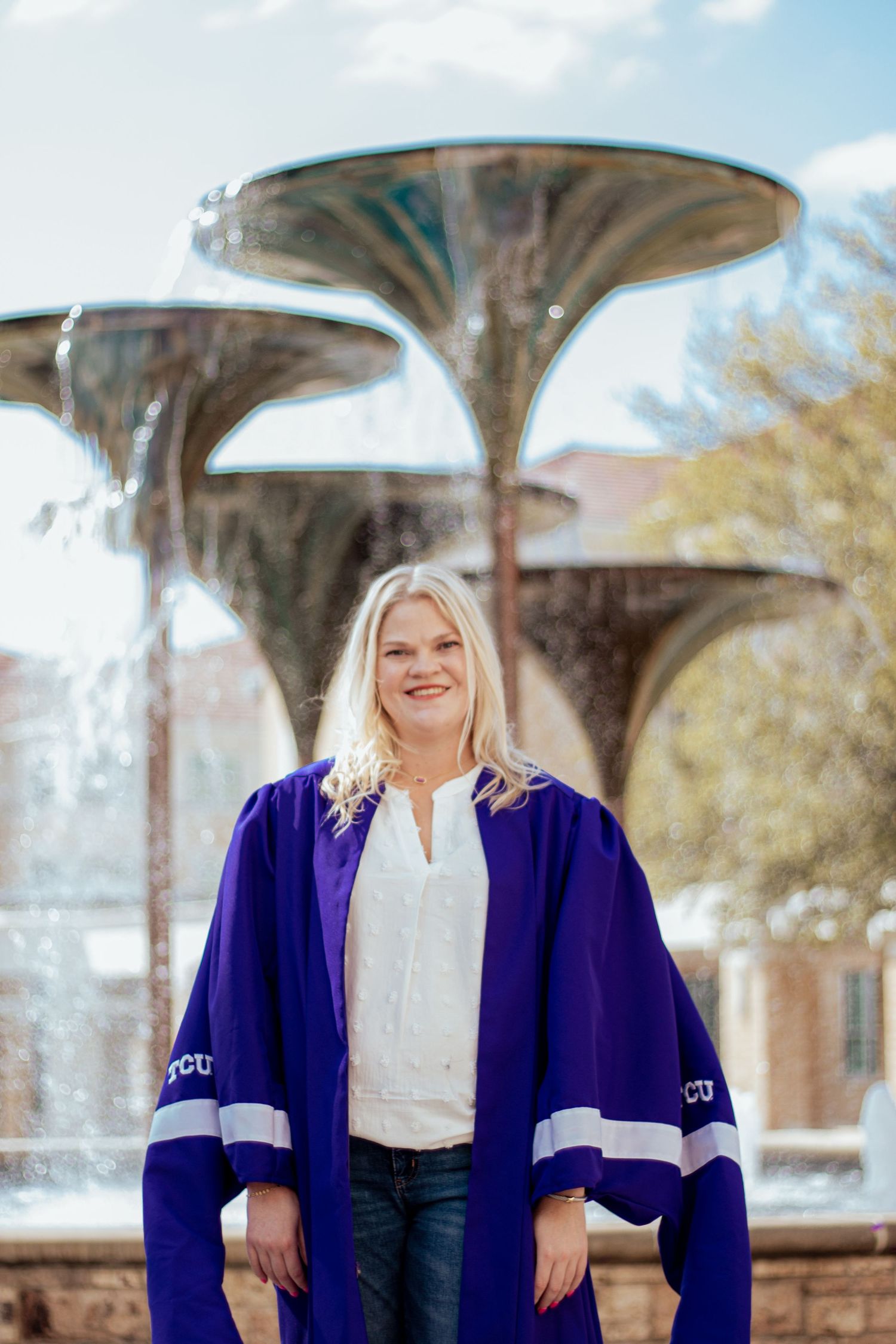 Graduate wearing purple academic regalia stands in front of decorative fountain on sunny campus day.