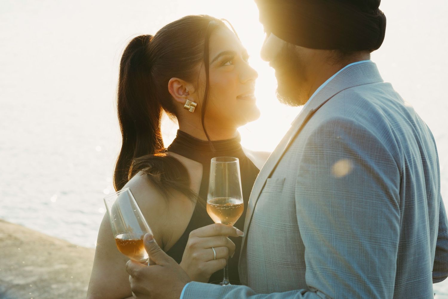 A romantic couple holds wine glasses at sunset with warm backlighting creating a dreamy atmosphere.