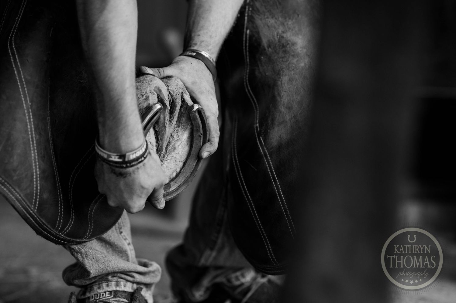 Black and white image of hands working with farrier tools on worn denim.