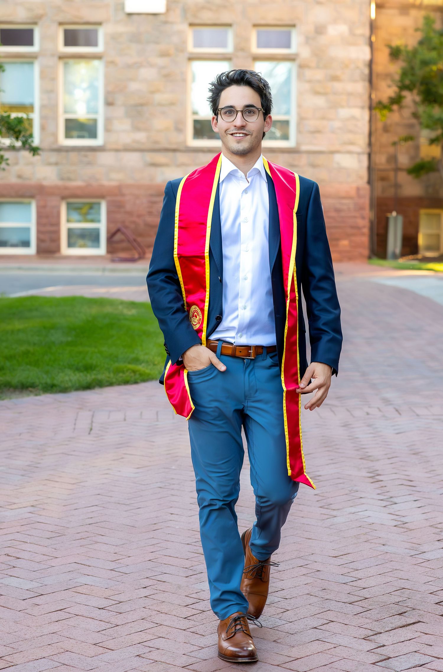 Graduate wearing navy suit with red and gold graduation stole walks along brick pathway on University of Denver campus.