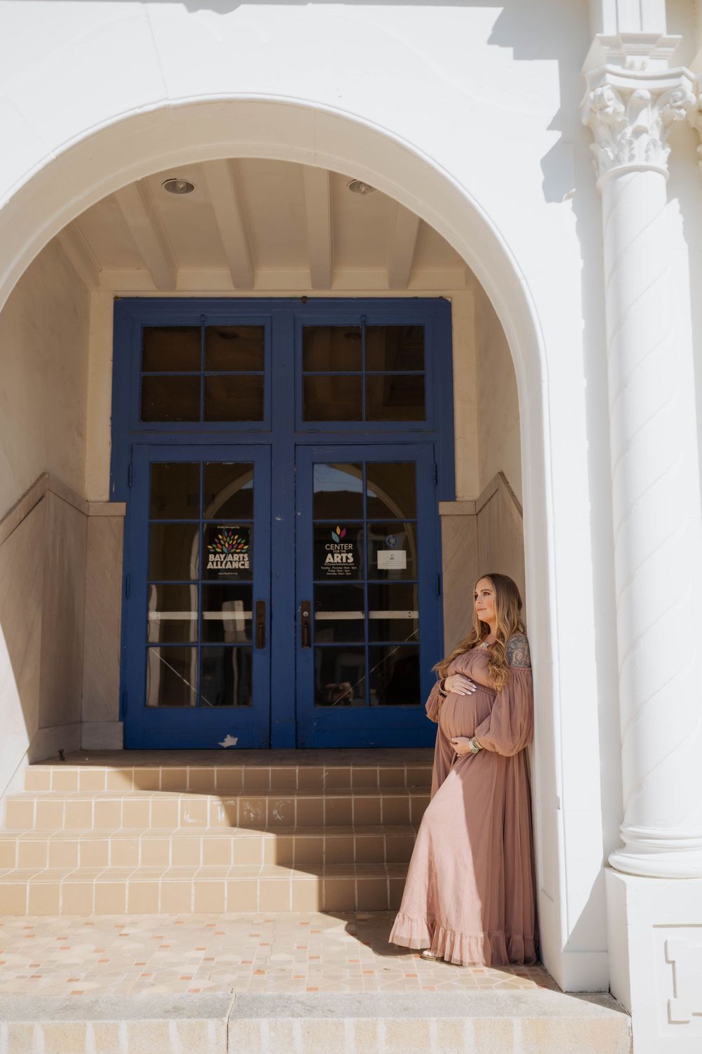 A person in a flowing mauve dress stands beneath an arched entrance with blue double doors and white columns.