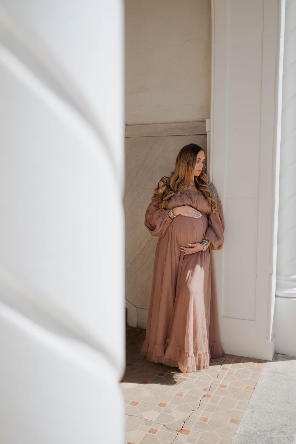 Expectant mother in a flowing mauve dress stands near white wall with natural window light creating soft shadows.