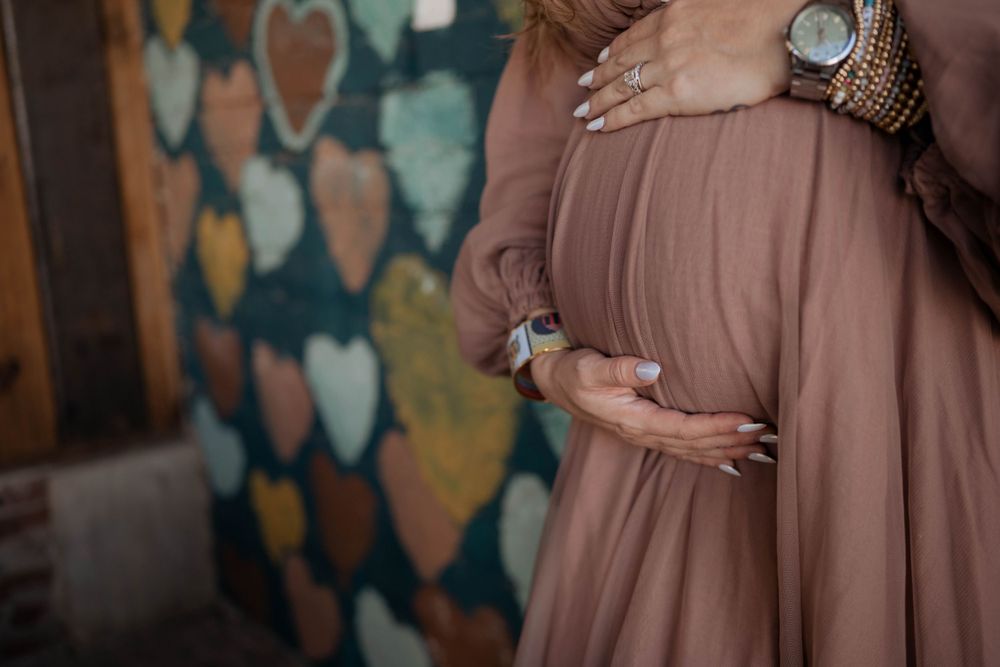 A pregnant woman in a dusty rose dress holds her baby bump while standing against a heart-patterned wall.