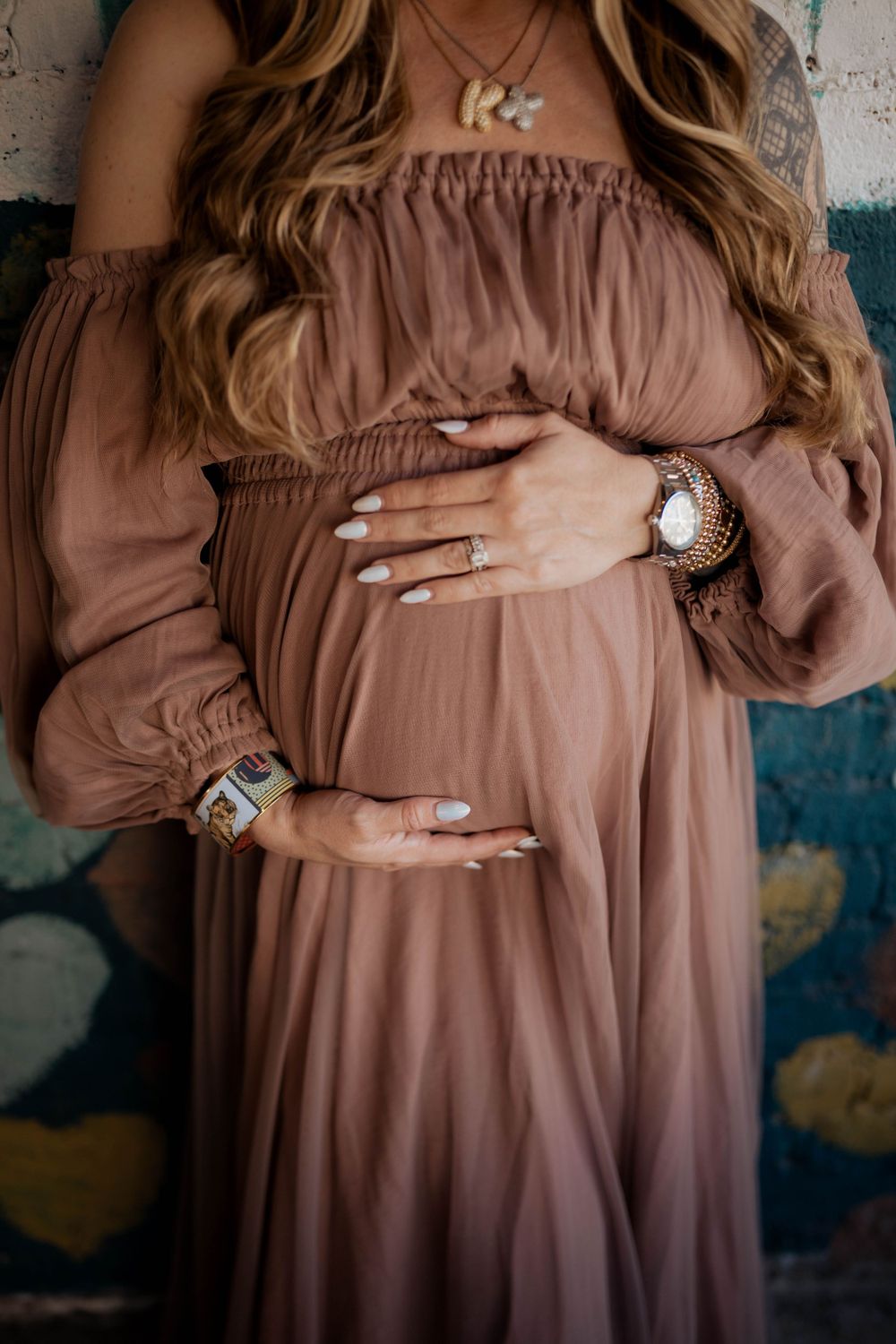 Close-up of hands adorned with rings resting on a flowing mauve maternity dress against a textured background.