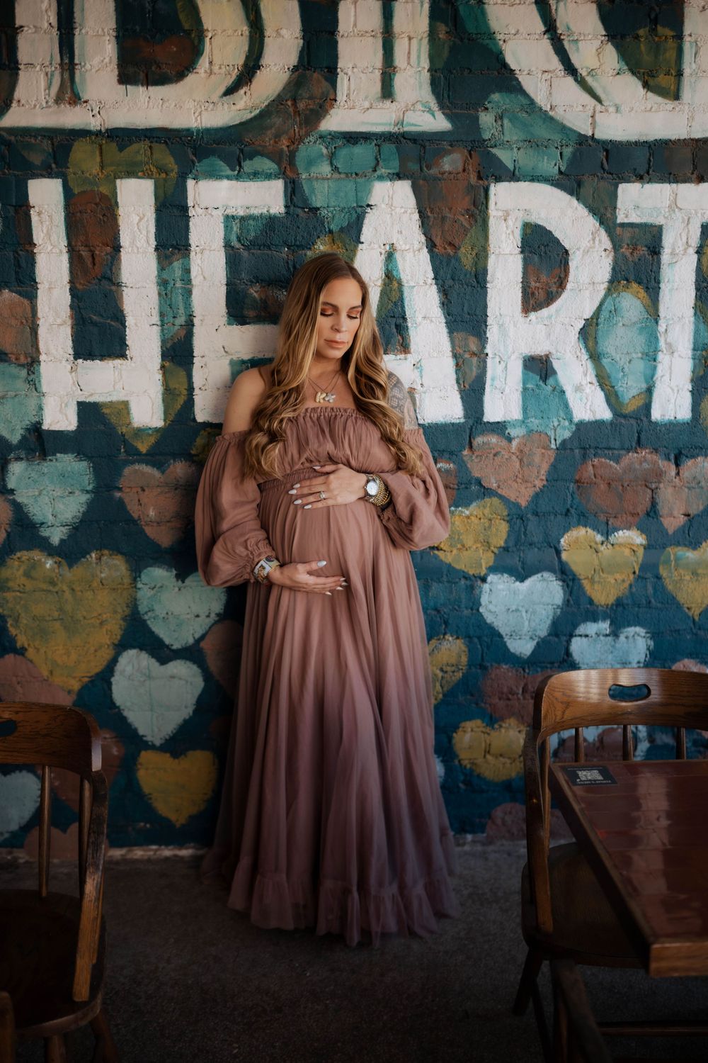 A maternity photo against a wall decorated with painted hearts, wearing a flowing mauve gown in a vintage setting.