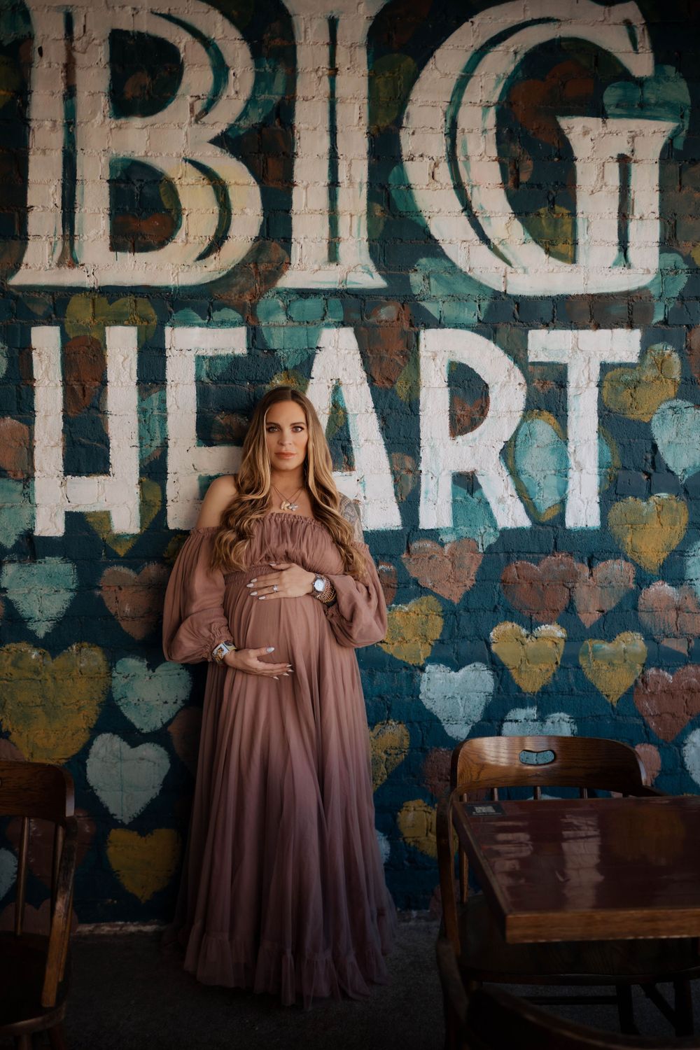 A maternity portrait in front of a rustic wall with blue hearts and text reading 'BIG HEART' painted on it.