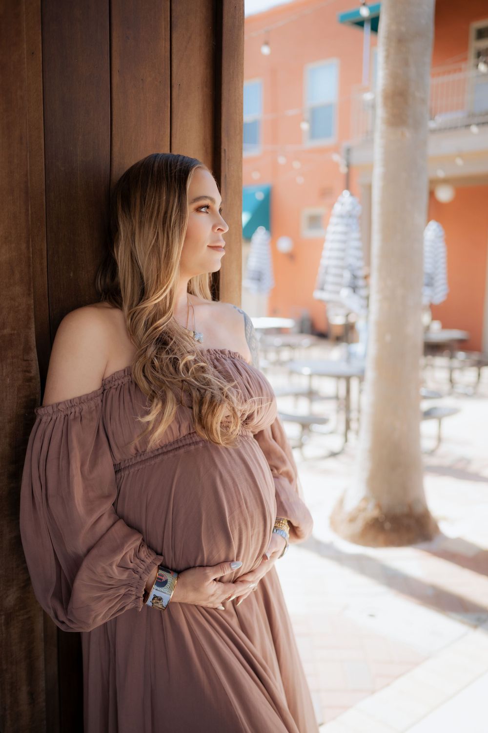 Expectant woman in mauve off-shoulder dress poses thoughtfully against a rustic wooden column with orange walls beyond.