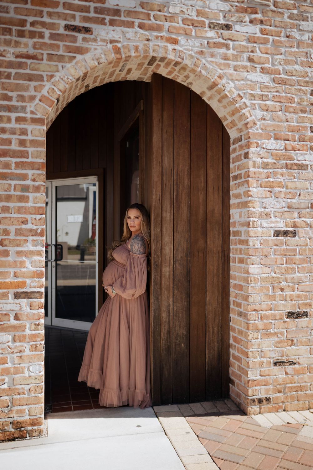 An expectant mother poses in a dusty rose gown under a brick archway with wooden doors.