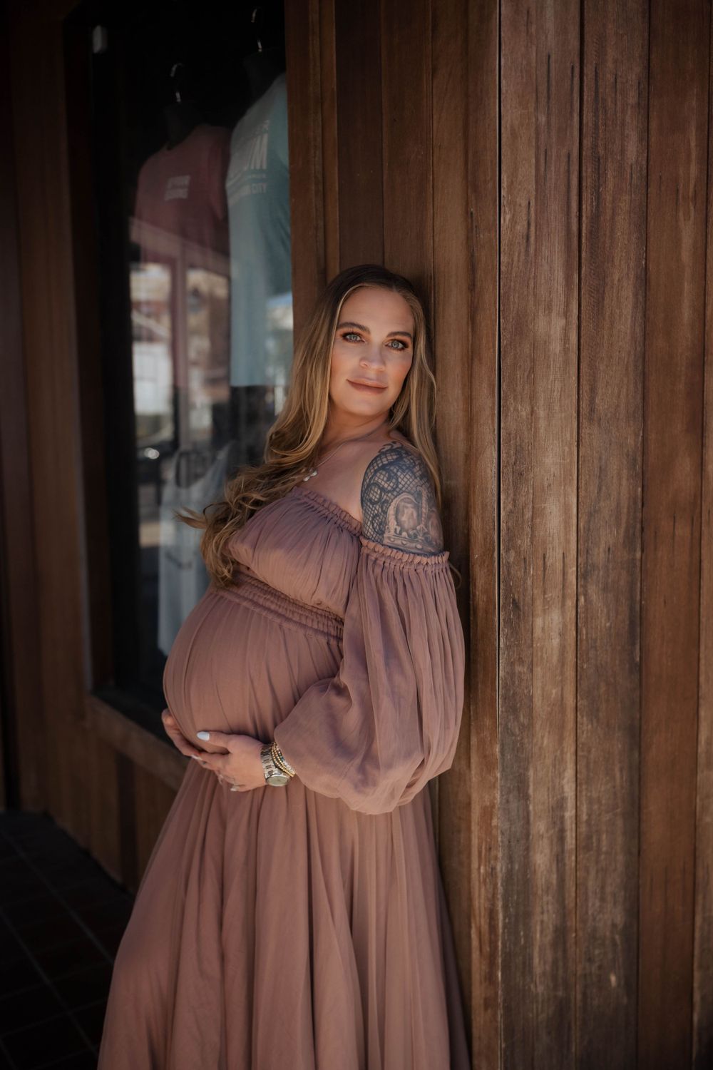 Expecting person in flowing dusty rose colored maternity dress poses by wooden doorway.