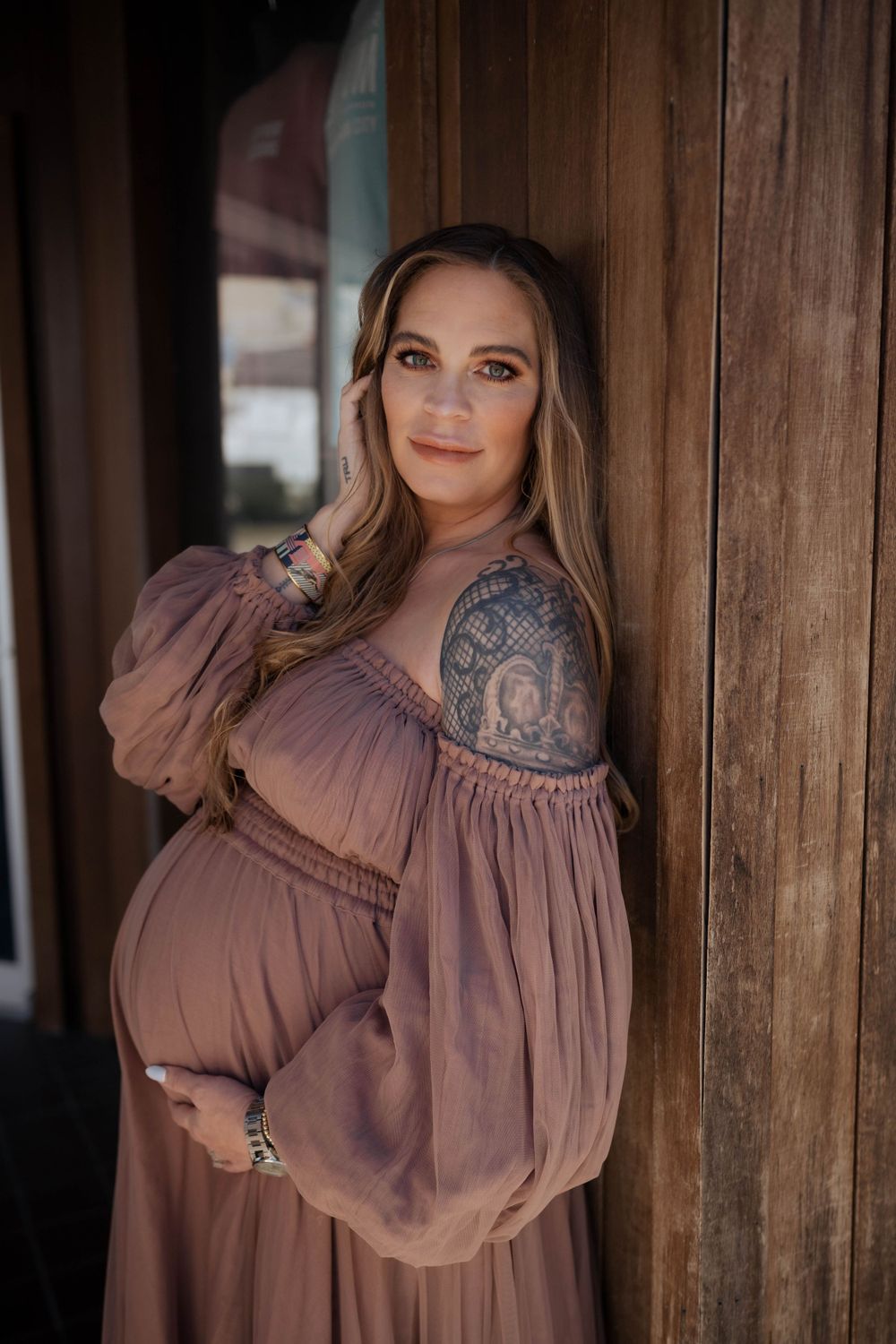 Expectant mother in flowing mauve dress with visible shoulder tattoo poses against wooden pillar in warm lighting.