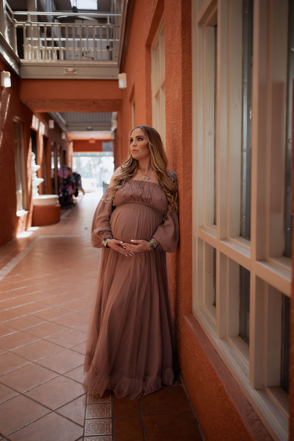 A maternity portrait in a coral-colored hallway featuring a flowing mauve dress and natural lighting.