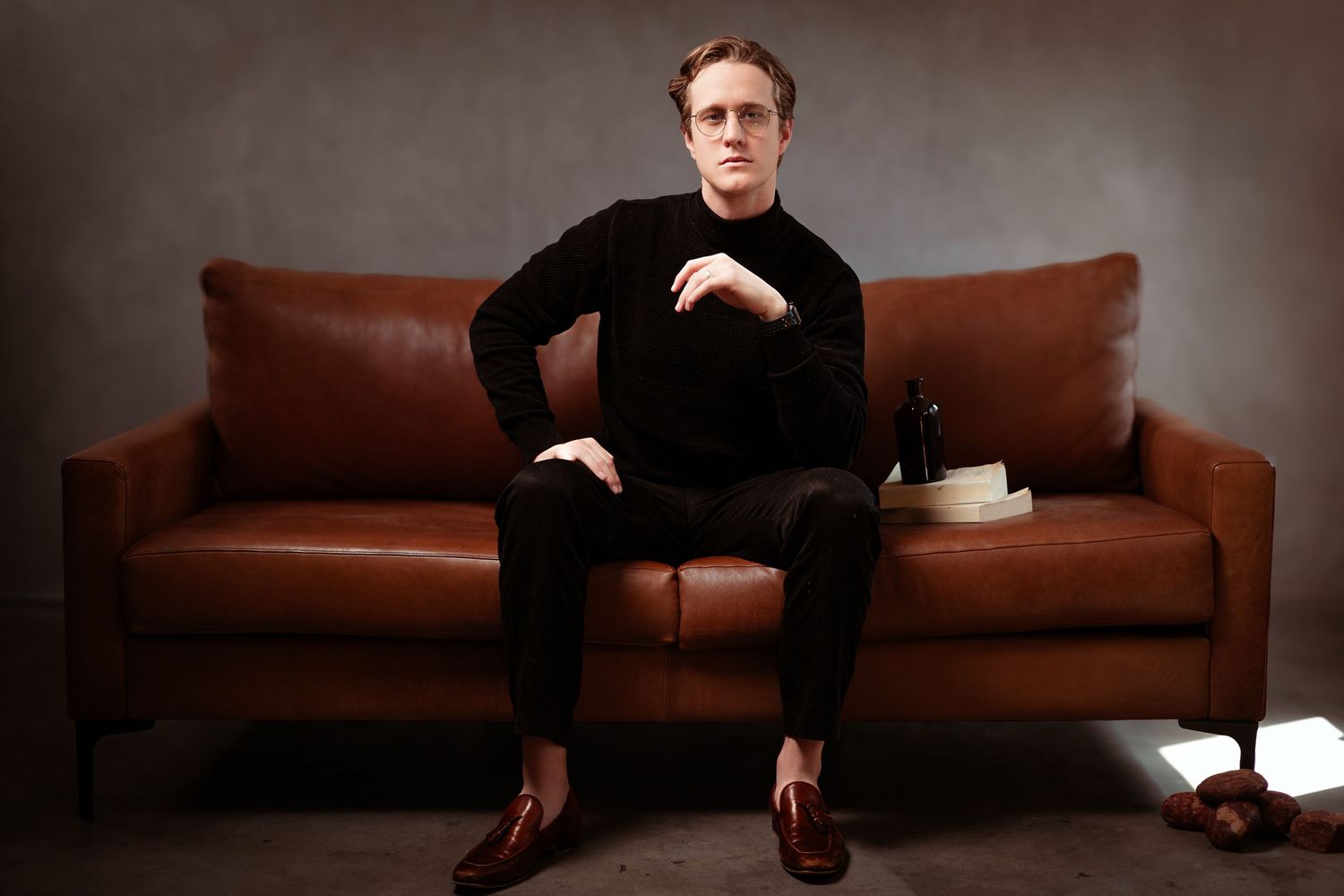 Person in all black attire sits on a brown leather couch against dark moody backdrop in studio setting.