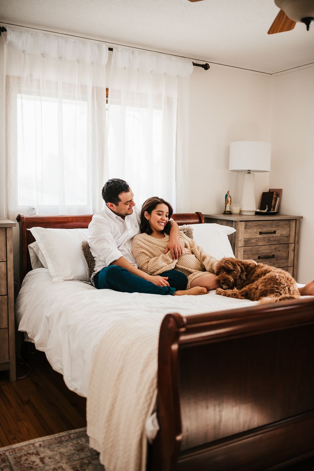 Couple relaxing with their dog on a sleigh bed in a cozy bedroom with white curtains and rustic furniture.