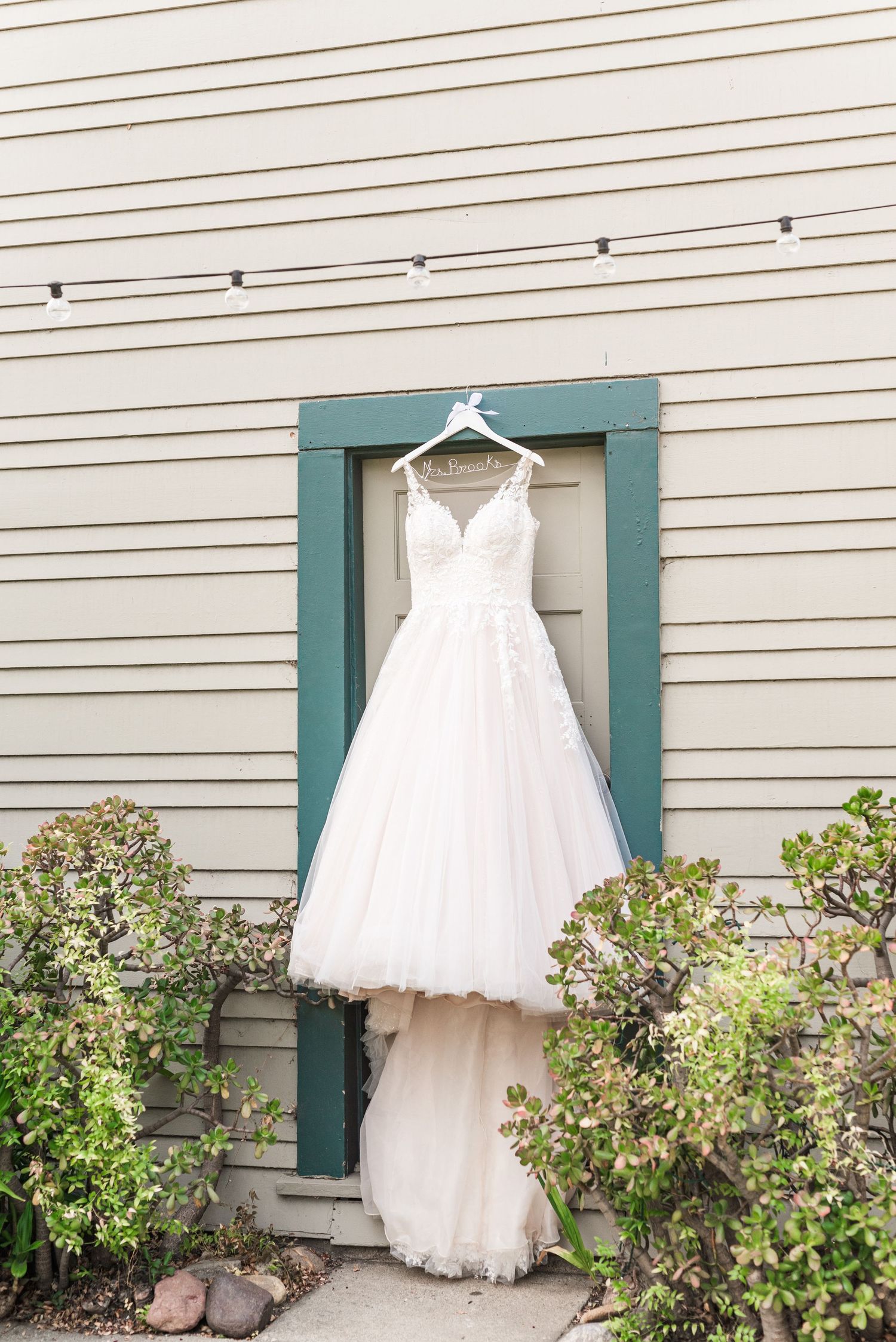 A white tulle wedding dress hangs on display against a green-trimmed window frame.