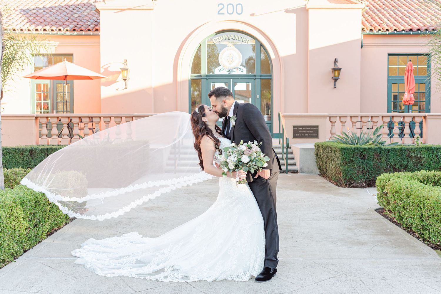 A dramatic wedding dip pose in front of a pink Spanish-style building with landscaped gardens.
