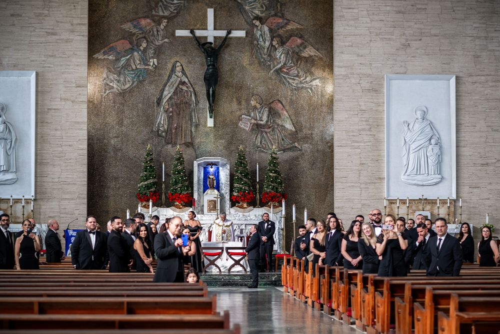 A church interior with wedding guests in dark attire gathered beneath a large crucifix and religious artwork.