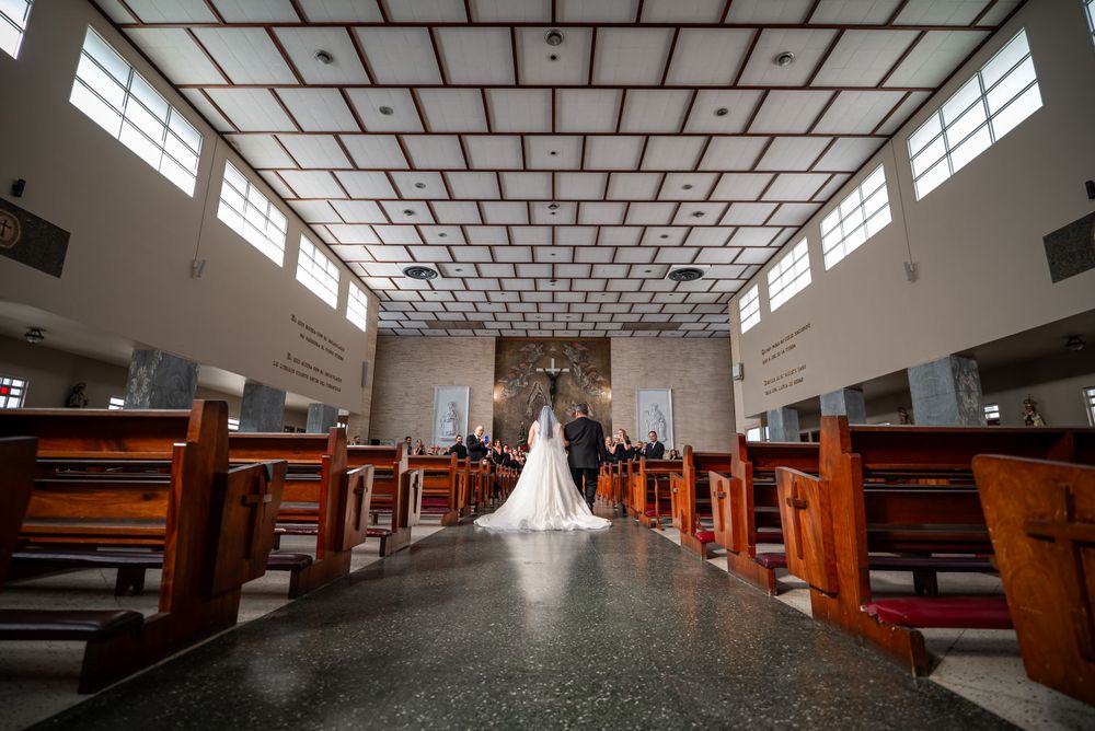 A long view down a church aisle with wooden pews and geometric ceiling tiles during a wedding ceremony.
