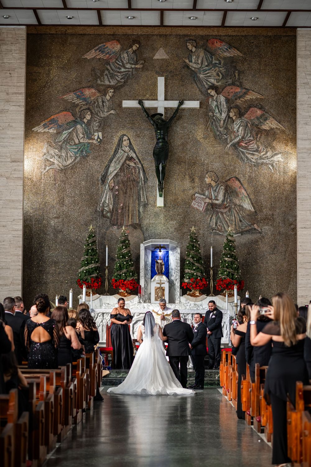 A wedding ceremony in a church with angel murals, poinsettias, and Christmas trees decorating the altar.