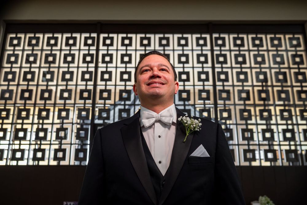 A formal portrait in front of a geometric patterned wall showing someone wearing a tuxedo with a boutonniere.