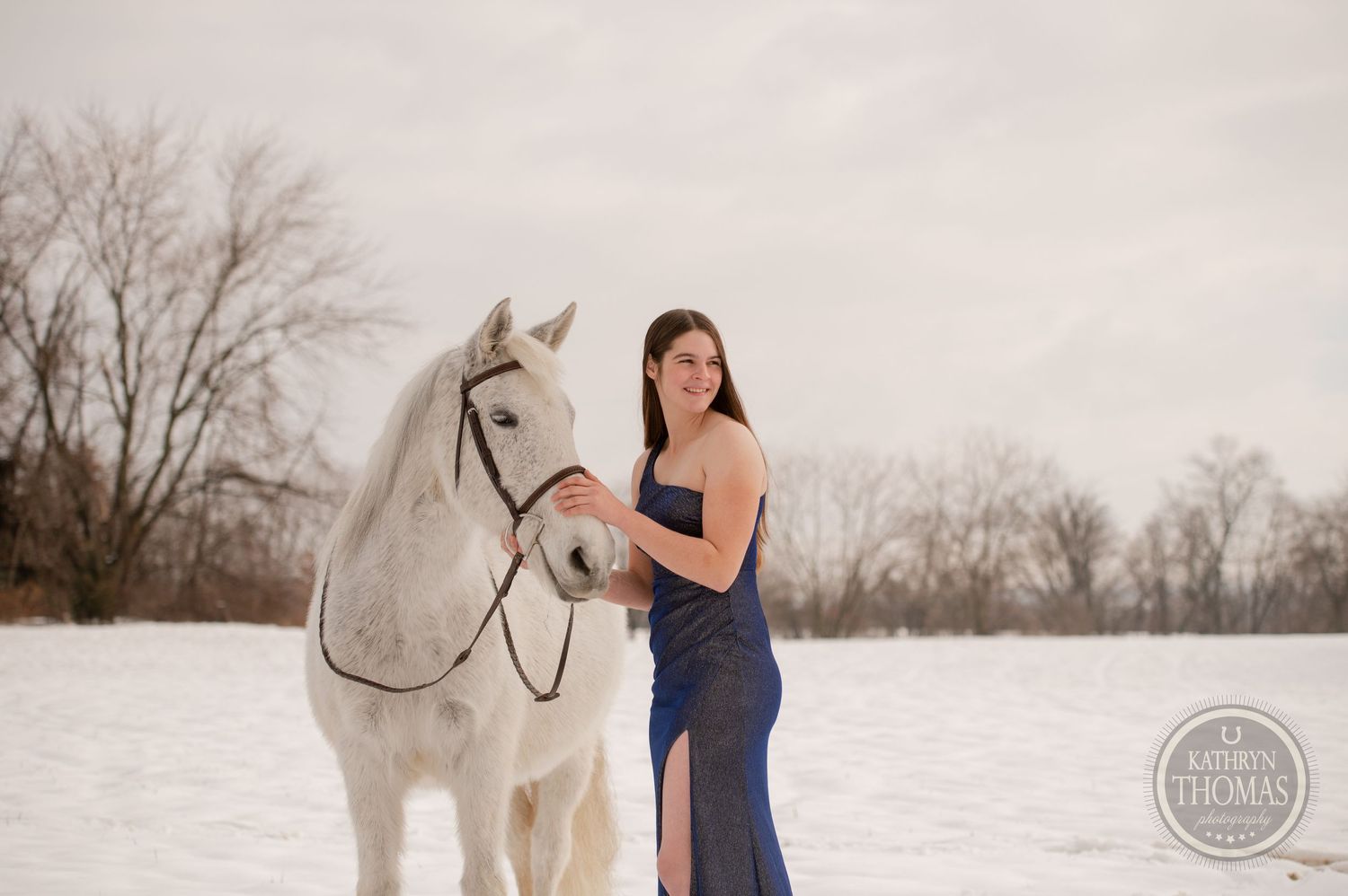 A white horse and person in a navy dress stand together in a serene snowy landscape.