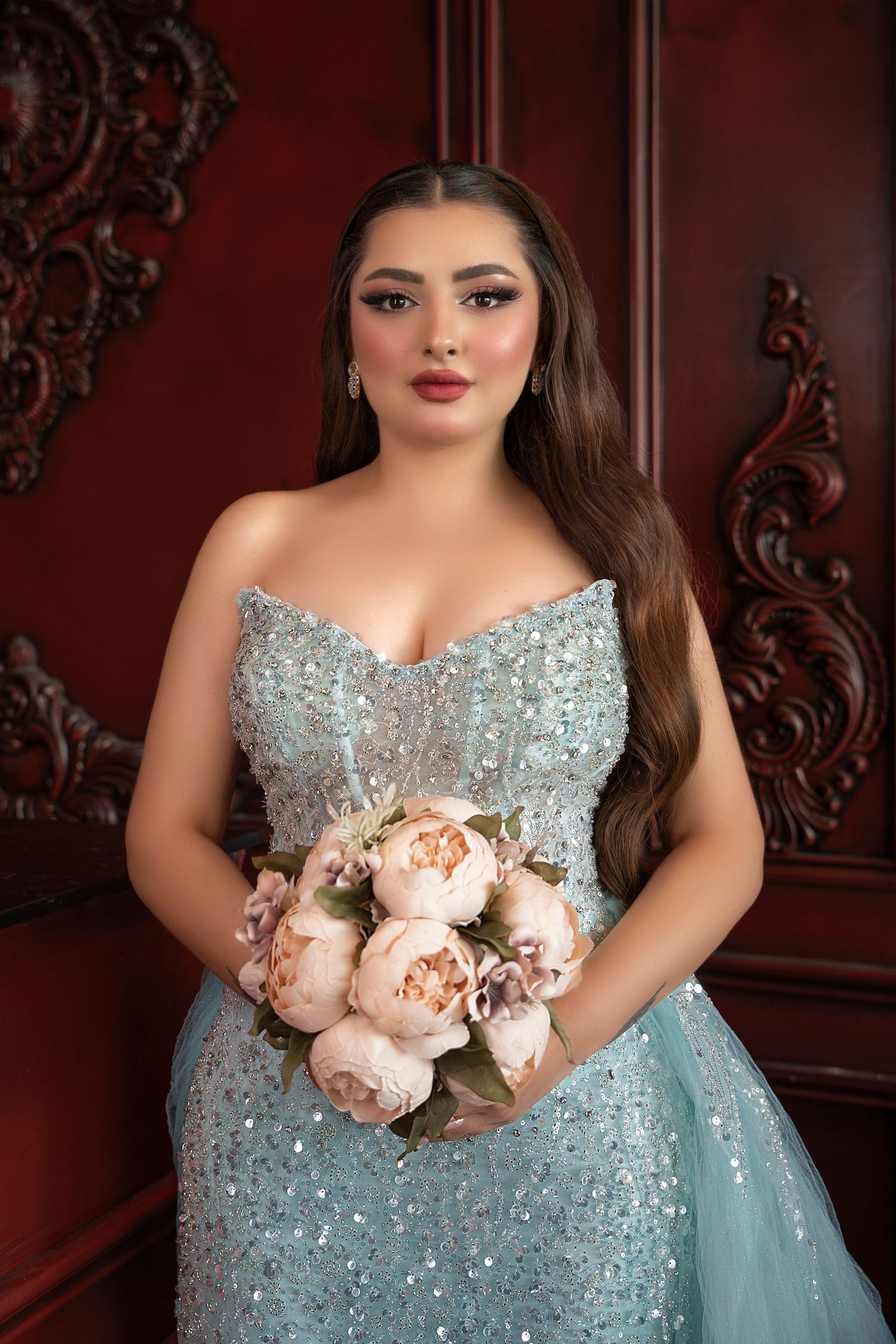 A formal portrait in a sparkling light blue gown holding a bouquet of pink peonies against an ornate dark red background.