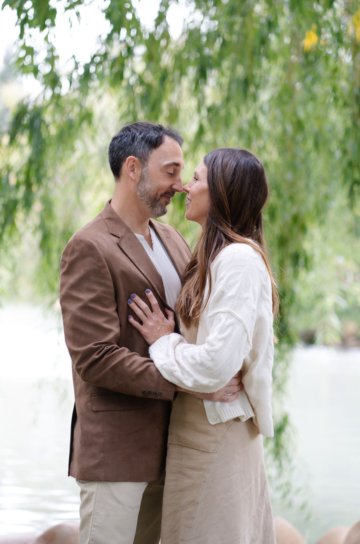 A romantic moment between a couple in brown and cream attire embracing under a willow tree.