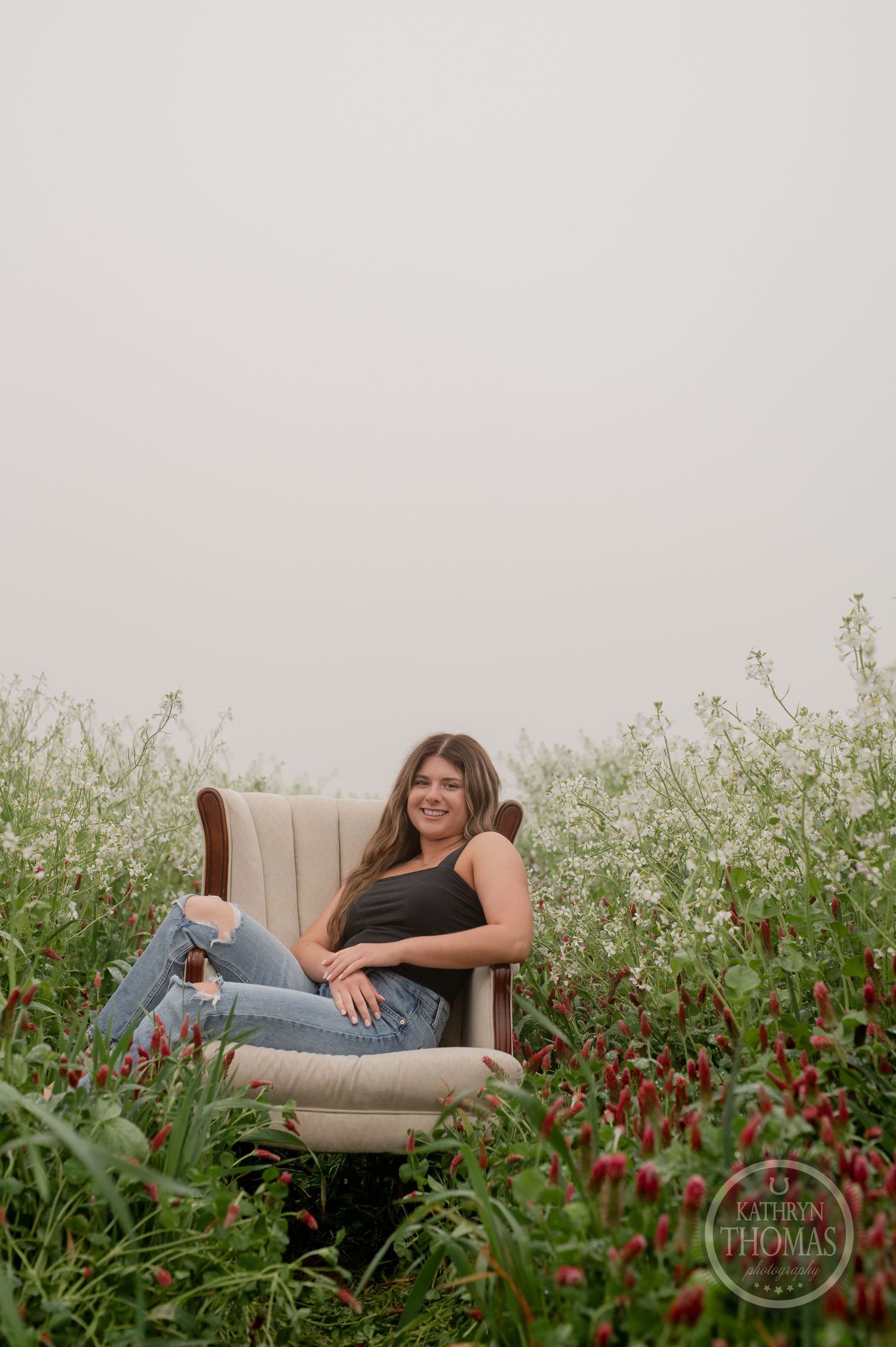 A relaxed portrait in distressed jeans and black tank top on a beige armchair among flowering plants.