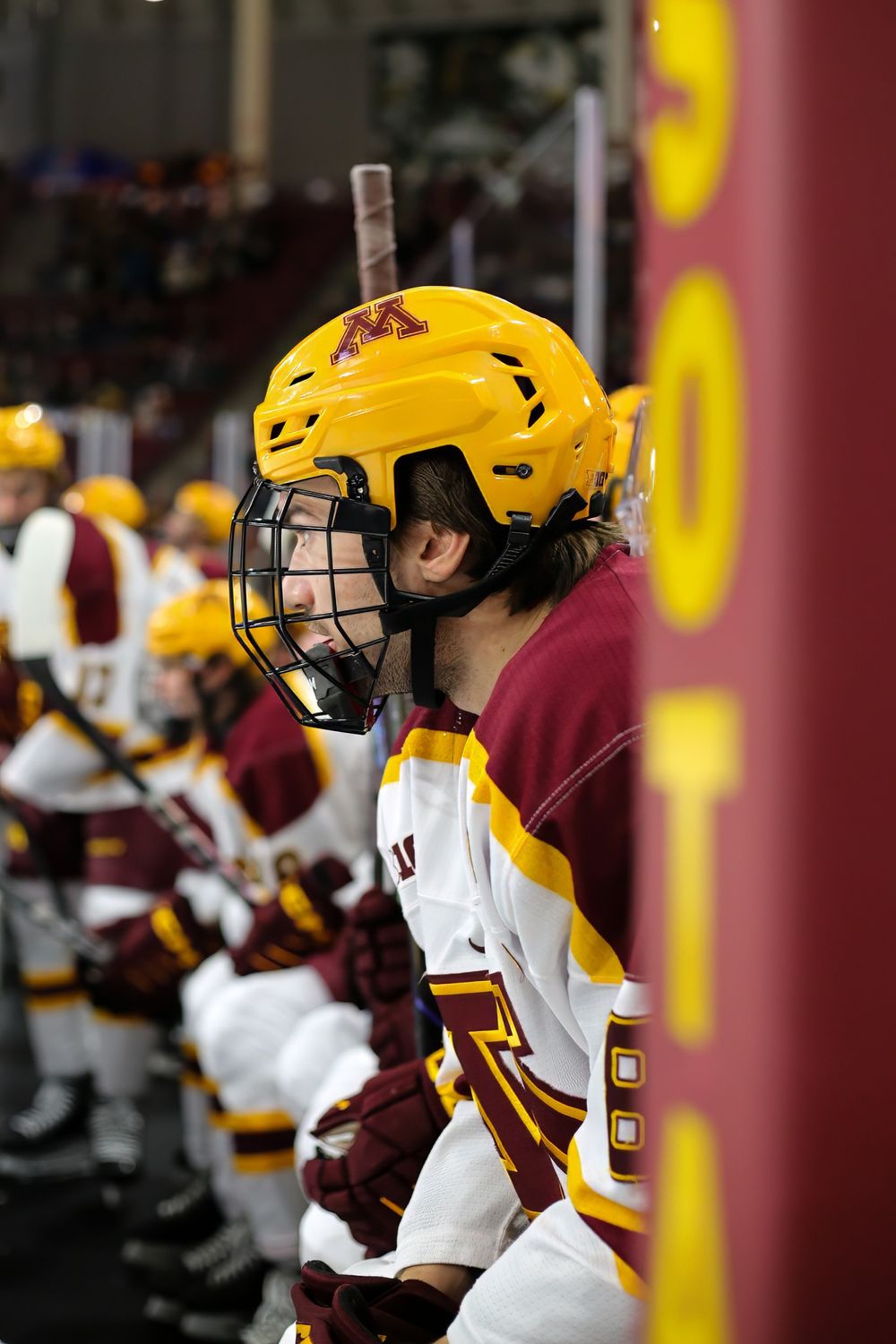 Hockey player in yellow helmet and maroon jersey sitting on bench during game.