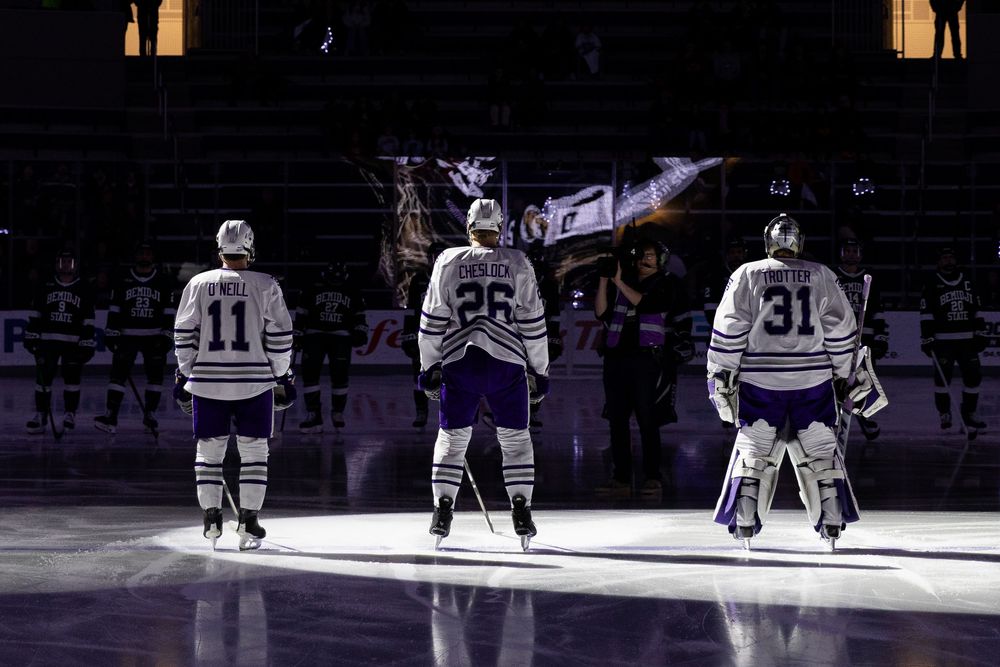 Hockey players stand on ice during pre-game ceremony with dramatic lighting and shadows in an arena setting.