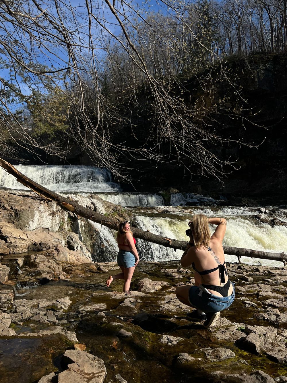 People climbing on rocky terrain near a cascading waterfall on a sunny day.
