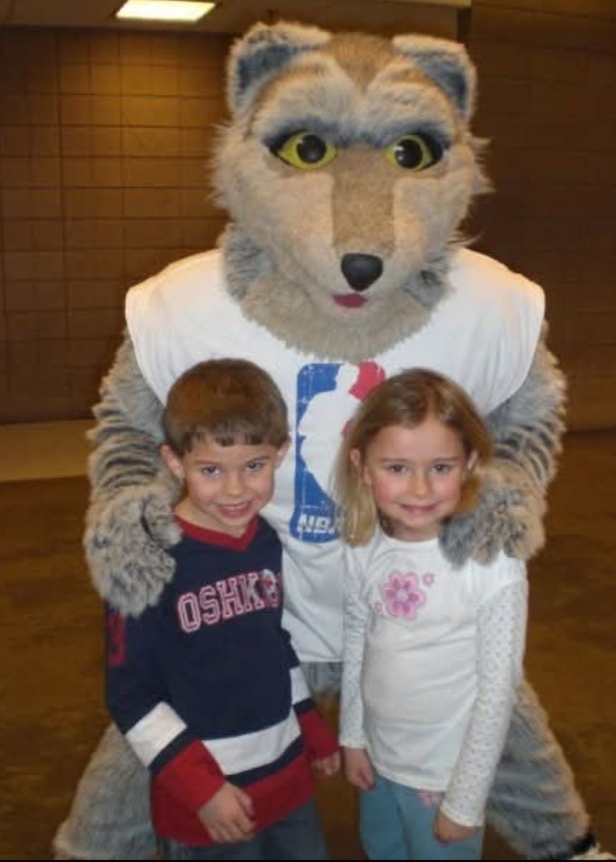 Large gray wolf mascot poses with two young children in an indoor setting.