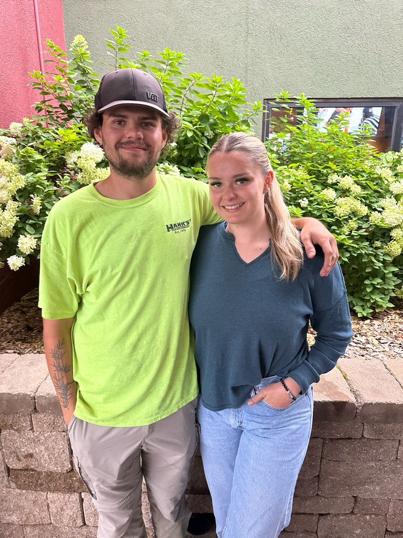 Casual couple standing together against greenery and brick wall background.